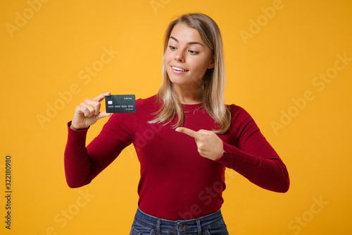 Amazed young blonde woman girl in casual clothes posing isolated on yellow orange background studio portrait. People lifestyle concept. Mock up copy space. Pointing index finger on credit bank card.