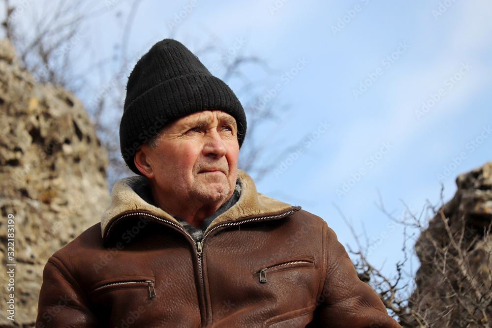 Brutal elderly man in leather coat in and knitted hat in a mountains on blue sky background. Concept of harsh highlander, travel and freedom, healthy old age and adventure on nature Brutal elderly man in leather coat in and knitted hat in a mountains on blue sky background. Concept of harsh highlander, travel and freedom, healthy old age and adventure on nature
