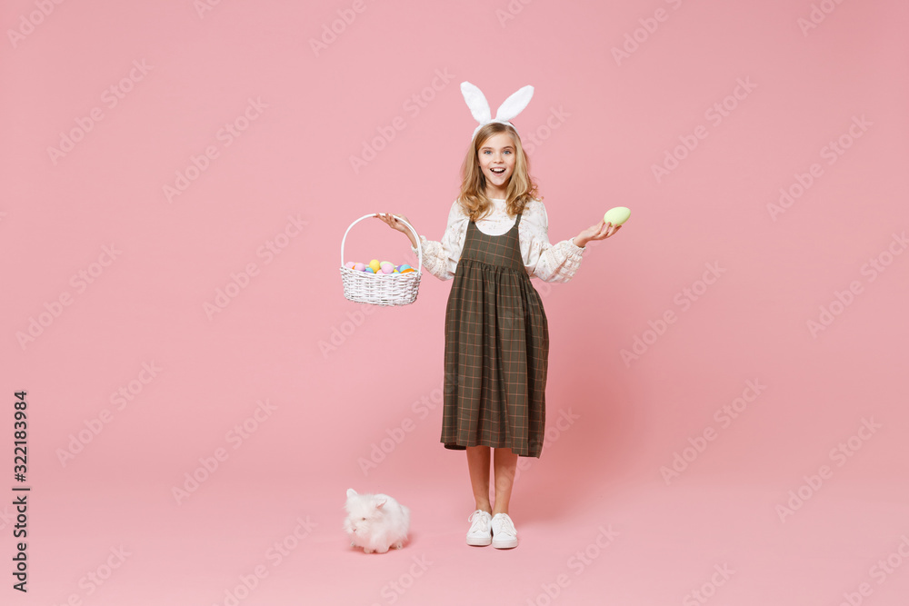 Fototapeta premium Little pretty blonde kid girl 11-12 years old in light spring dress hold fluffy white bunny rabbit, wicker basket with eggs isolated on pastel pink background. Childhood lifestyle Happy Easter concept