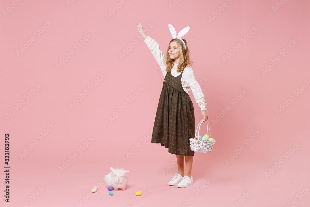 Little pretty blonde kid girl 11-12 years old in light spring dress hold fluffy white bunny rabbit, wicker basket with eggs isolated on pastel pink background. Childhood lifestyle Happy Easter concept