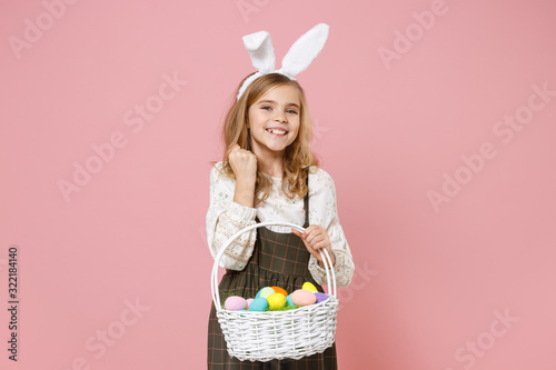 Little pretty blonde kid girl 11-12 years old in spring dress, bunny rabbit ears hold in hand wicker basket colorful eggs isolated on pastel pink background. Childhood lifestyle Happy Easter concept.