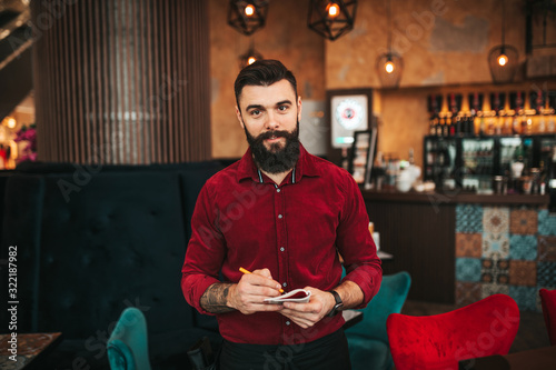Young handsome bearded barista in red shirt working in modern cafe bar.