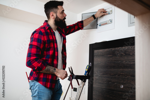 Canvas Print Young bearded repairman standing on ladder and repairing fuse box