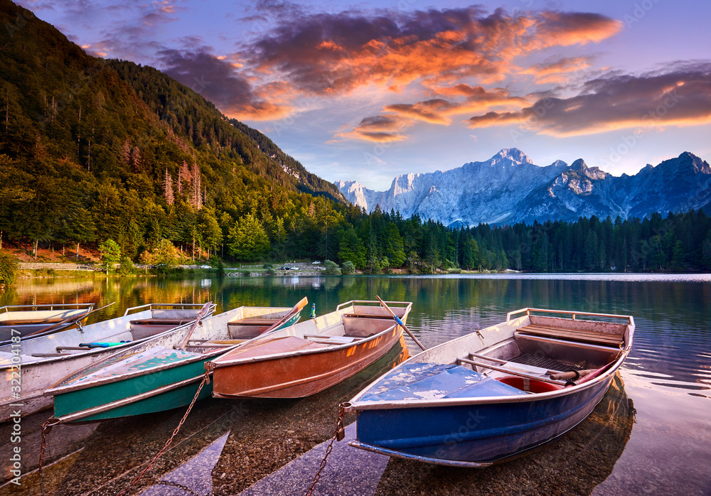Amazing view on Lago di Fusine Inferiore at sunrise. Splendid morning scene of Julian Alps, Province of Udine, Italy, Europe. Beautiful forests are reflected in the quiet lake. Four pleasure boats
