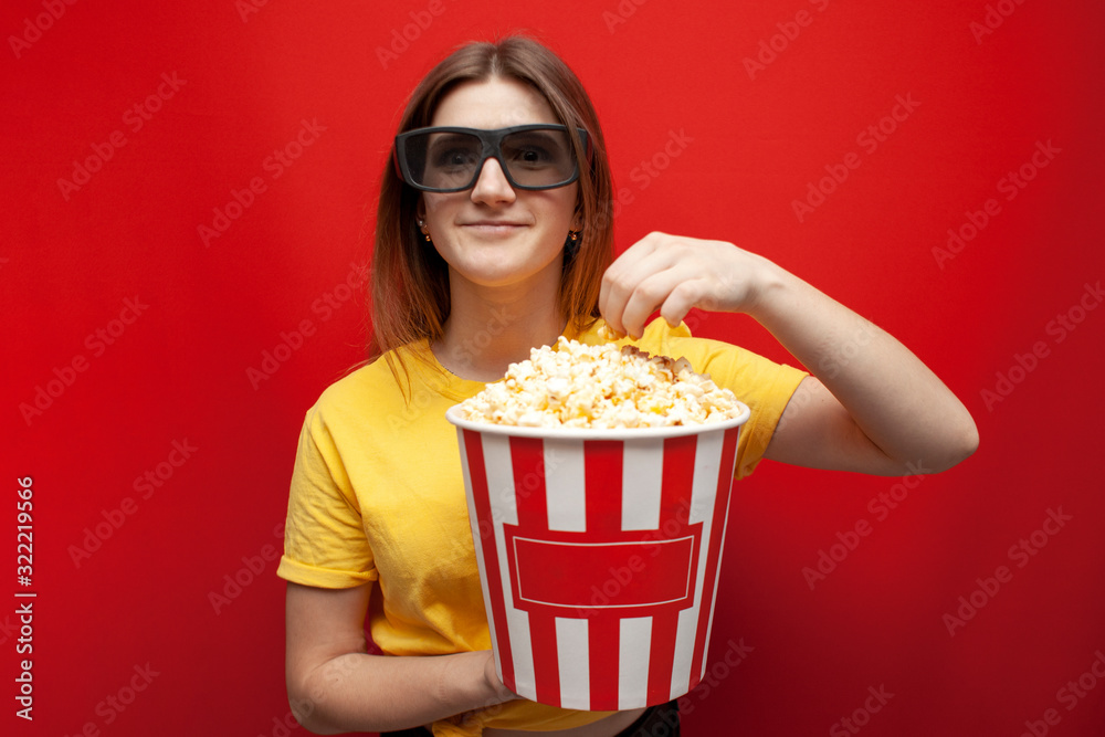 beautiful young girl in 3D glasses eating popcorn and watching a movie on a red color background