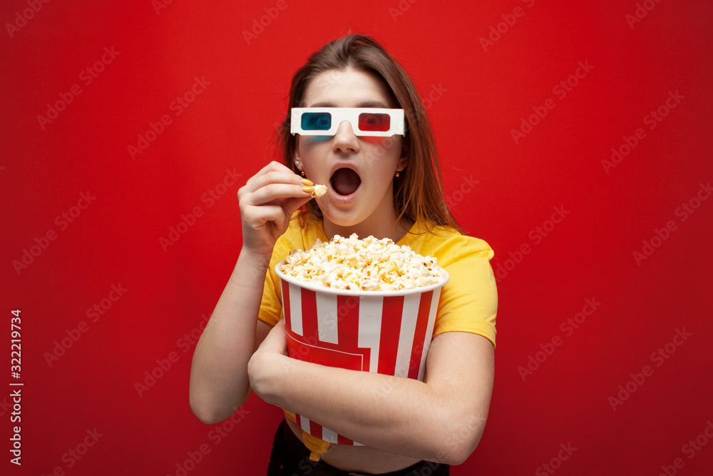 beautiful young emotional girl in 3D glasses eats popcorn, watches a movie and screams on a red color background, surprised viewer beautiful young emotional girl in 3D glasses eats popcorn, watches a movie and screams on a red color background, surprised viewer