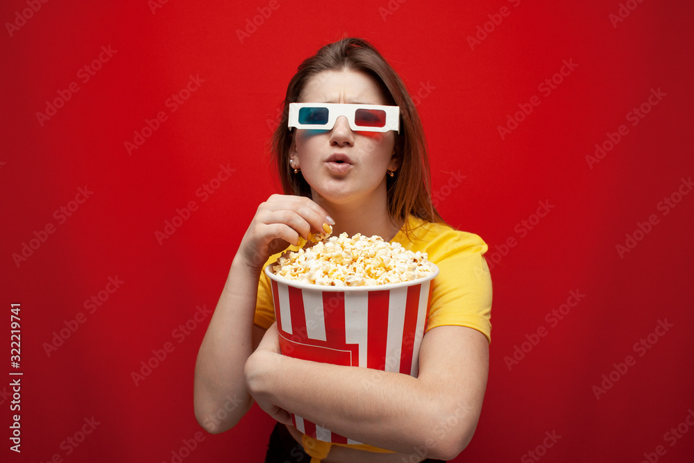 beautiful young emotional girl in 3D glasses eats popcorn, watches a movie and screams on a red color background, surprised viewer beautiful young emotional girl in 3D glasses eats popcorn, watches a movie and screams on a red color background, surprised viewer