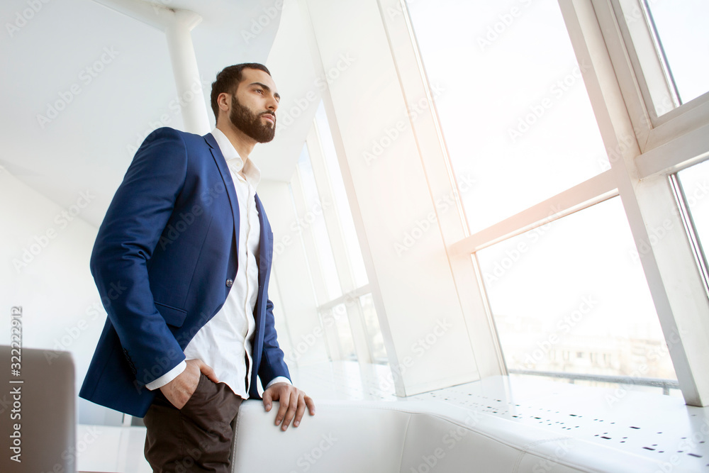 successful handsome businessman in a blue suit stands against the background of a white office and looks out the window, portrait of a director at the workplace successful handsome businessman in a blue suit stands against the background of a white office and looks out the window, portrait of a director at the workplace