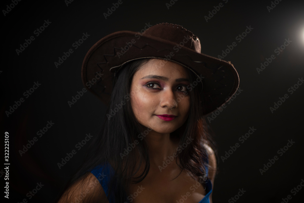 Fashion portrait of young and attractive Indian Bengali brunette girl with blue western dress with cowboy hat under spotlight in front of black studio background.Indian fashion portrait and lifestyle.