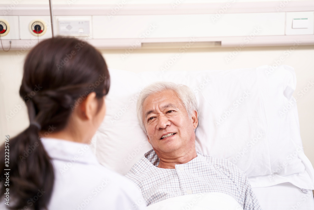 asian old man lying in bed talking to young female doctor in hospital ward