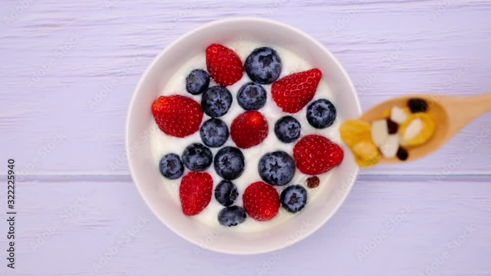A man using wooden spoon to add cereal, raisin, almond slices to a bowl of natural yogurt with fresh berries. A man using wooden spoon to add cereal, raisin, almond slices to a bowl of natural yogurt with fresh berries.