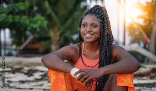 Charming beautiful young african american woman girl with black pigtails with piercings on her face, sitting on the beach during sunset