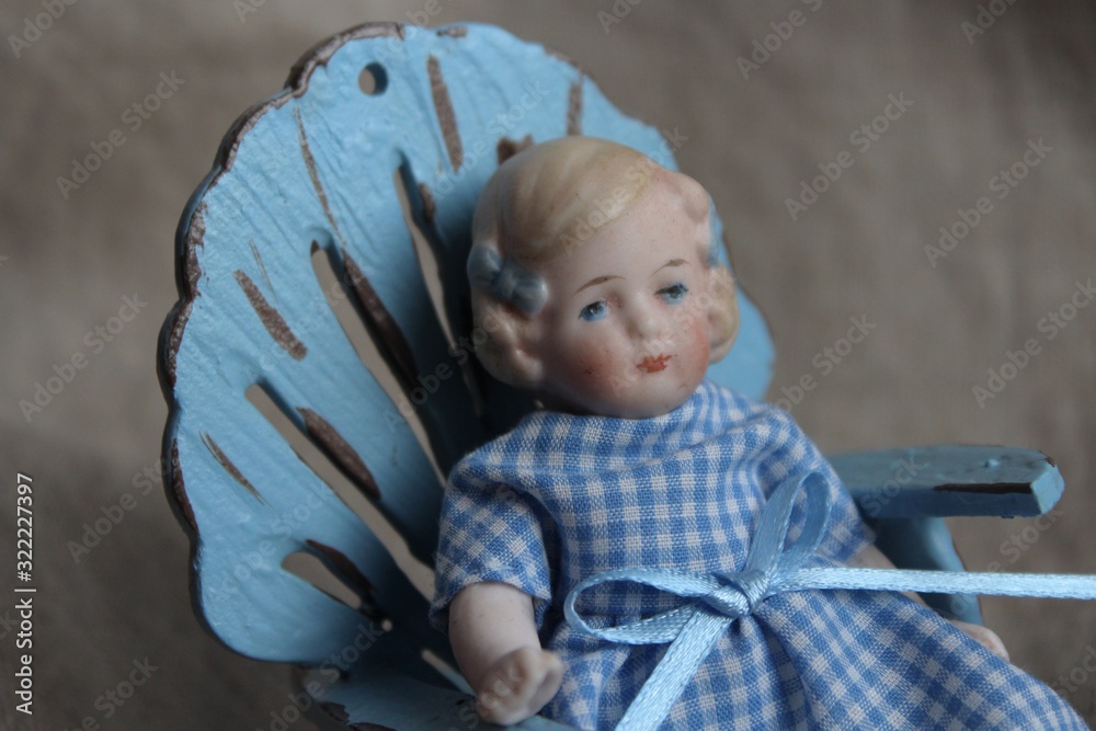 Φωτογραφία A little porcelain antique doll for a dollhouse, with two blue bows in blonde hair and checkered blue dress on a rough linen tissue background of natural color