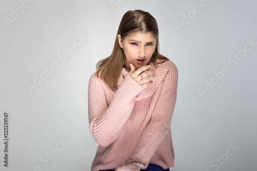 Close up portrait of a young Caucasian woman wearing a pink sweater on a grey background. She looks disgusted or repulsed by something