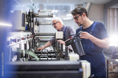 Serious young worker in glasses and overall adding ink into printing press at factory