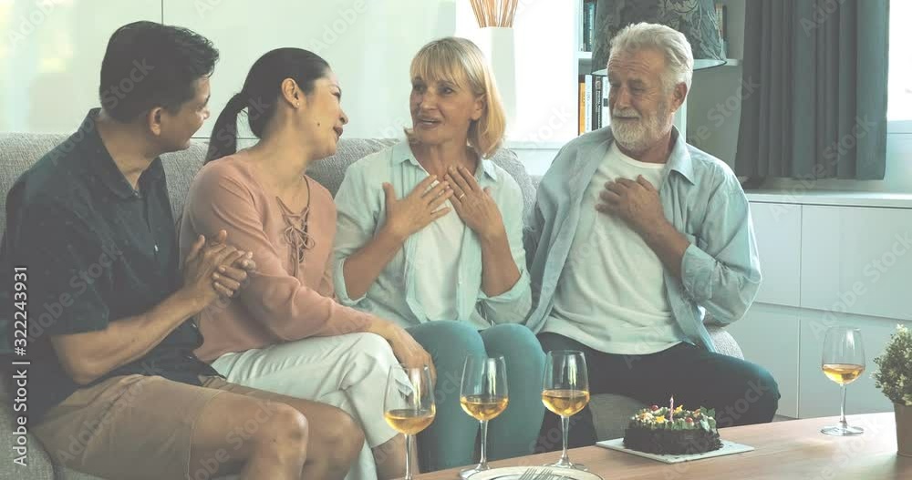 Senior couple hugging on birthday party with their friends in living room. Caucasian, Asian, Thai people sitting on couch at home. Retirement lifestyle concept. Senior couple hugging on birthday party with their friends in living room. Caucasian, Asian, Thai people sitting on couch at home. Retirement lifestyle concept.