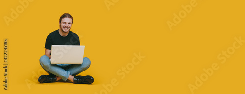 A happy boy with long hair and brown beard sitting with a computer on a yellow background with blank space