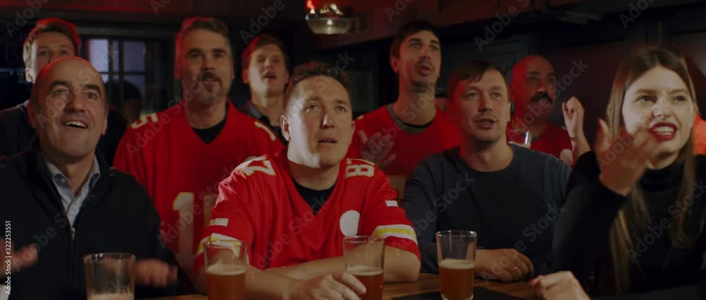 MED Model released, American football fans wearing generic red uniform watching a game on a TV in a sport pub, rooting for their team. 4K UHD, shot on ARRI Alexa Mini with Cooke S4 lenses