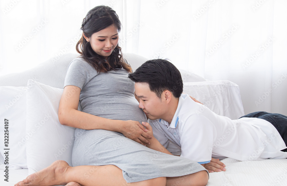 Beautiful Asian pregnancy woman, husband hold belly, tummy with love, kiss, caring togetherness on bed at home, excited motherhood awaiting for childbirth sharing moment with couple, family concept Beautiful Asian pregnancy woman, husband hold belly, tummy with love, kiss, caring togetherness on bed at home, excited motherhood awaiting for childbirth sharing moment with couple, family concept