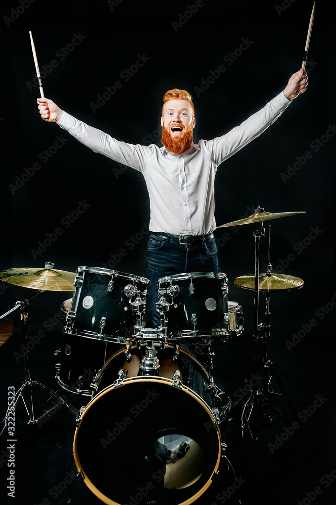 Fototapeta premium Portrait of a red-haired emotional man playing drums and cymbals and holding a stick. isolated on a dark background