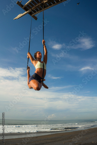 aerial yoga beach workout - young attractive and healthy woman practicing aero-yoga training balance body and mind control hanging from ropes above the sea