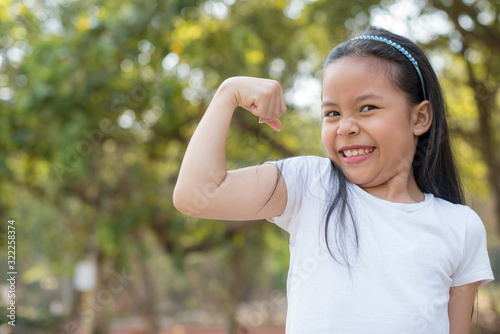 happy Little asian girl child standing showing front teeth with big smile. showing arms muscles smiling proud. Looking camera showing biceps. fresh healthy green bio background. Fitness concept.