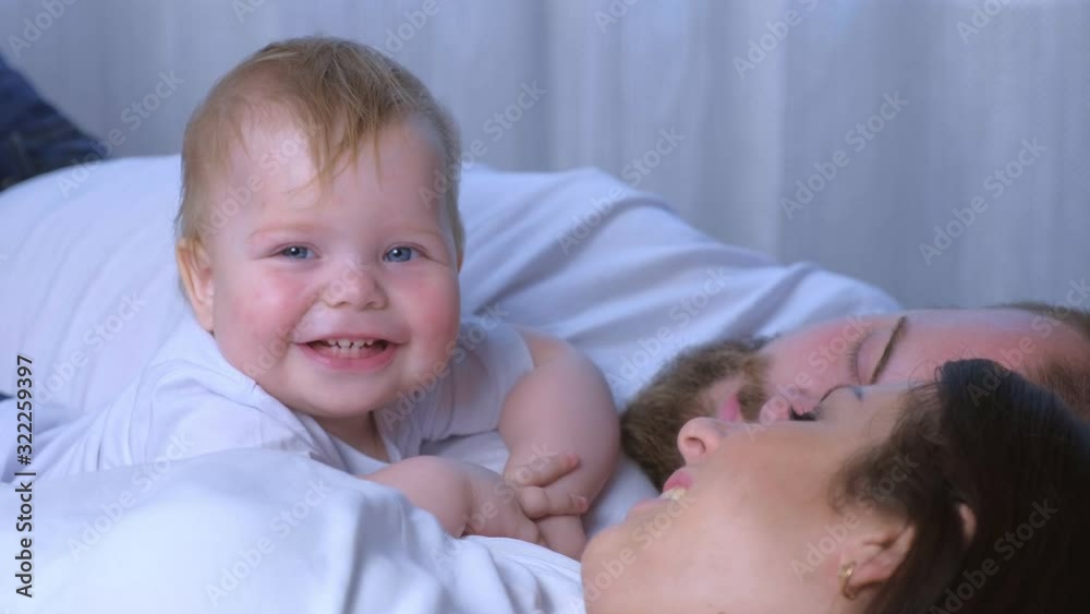 Portrait of smiling one year old baby looking at camera lying with parents at home. Family lovely moments, family look style. Mom, dad and baby are resting together, happy relationships. Portrait of smiling one year old baby looking at camera lying with parents at home. Family lovely moments, family look style. Mom, dad and baby are resting together, happy relationships.