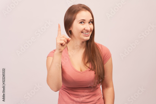 She found answer. Studio shot of beautiful creative european woman raising index finger up and smiling broadly, being in great mood after making up great plan or suggestion, happy to share it.