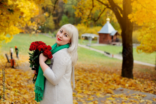 An adult woman of middle age  large size  stands and smiles with red roses in her hands in nature  against the background of yellow autumn trees. She looks over her shoulder at the camera and smiles.