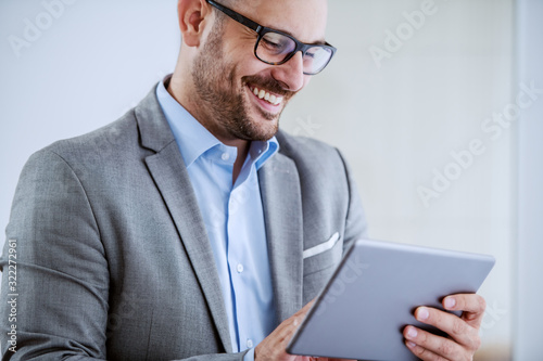 Close up of cheerful caucasian classy businessman in suit and with eyeglasses standing next to window and using tablet at his office.