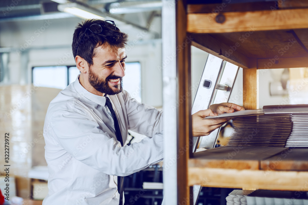 Fototapeta premium Smiling caucasian bearded supervisor in shirt and tie taking offset printing plate while standing in printing shop.