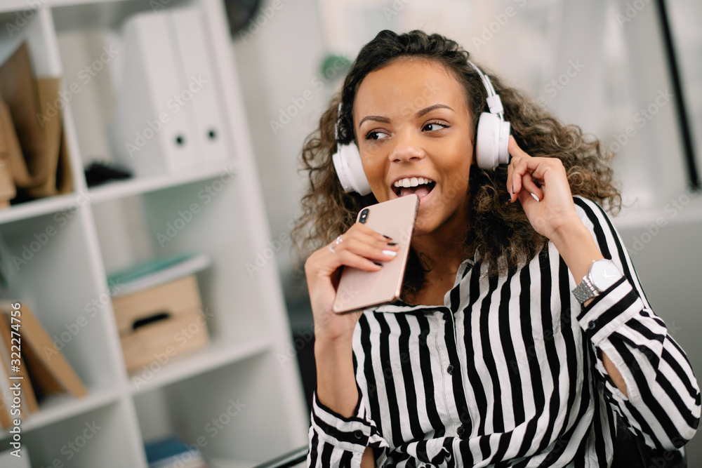 Businesswoman enjoying at work. Young african woman singing while listening music on phone in office. Businesswoman enjoying at work. Young african woman singing while listening music on phone in office.