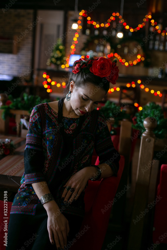 vertical photo of a girl in bright clothes and flowers in her hair, who sits sadly with her head bowed in the background of a beautiful interior