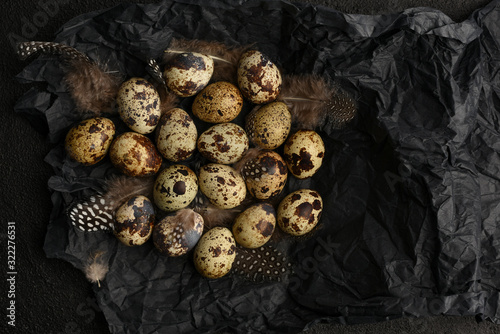 Quail eggs and feathers on black crumpled wrapping paper. A lot of spotty little eggs on a black textured background. Copy space. Healthy food.