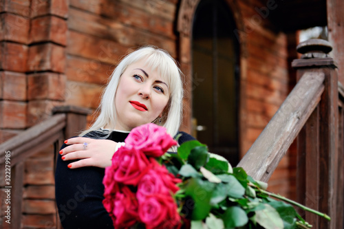 An adult plump blonde in a black dress stands with red roses in her hands, her head bowed and dreamily looking up, against the background of an old wooden house. Closeup. © alexbard