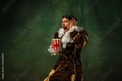 Emotional watching soccer. Portrait of medieval young man in vintage clothing standing on dark background. Male model as a duke, prince, royal person. Concept of comparison of eras, modern, fashion.