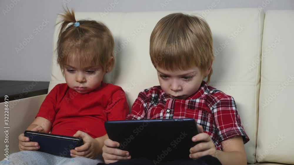 Children in red shirts are lying on a bright sofa playing on a tablet in a social network. Blond boy and girl watch videos on a digital tablet.