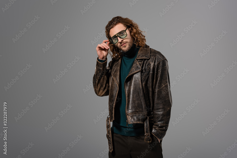 handsome man in leather jacket touching glasses and looking at camera isolated on grey handsome man in leather jacket touching glasses and looking at camera isolated on grey