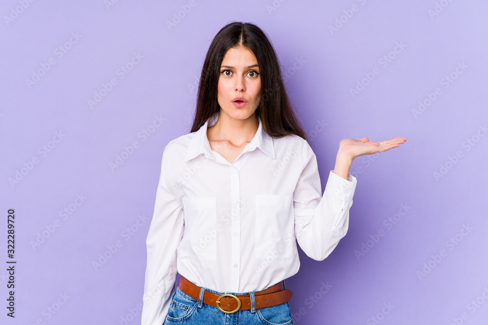 Young caucasian woman isolated on purple background impressed holding copy space on palm. Young caucasian woman isolated on purple background impressed holding copy space on palm.