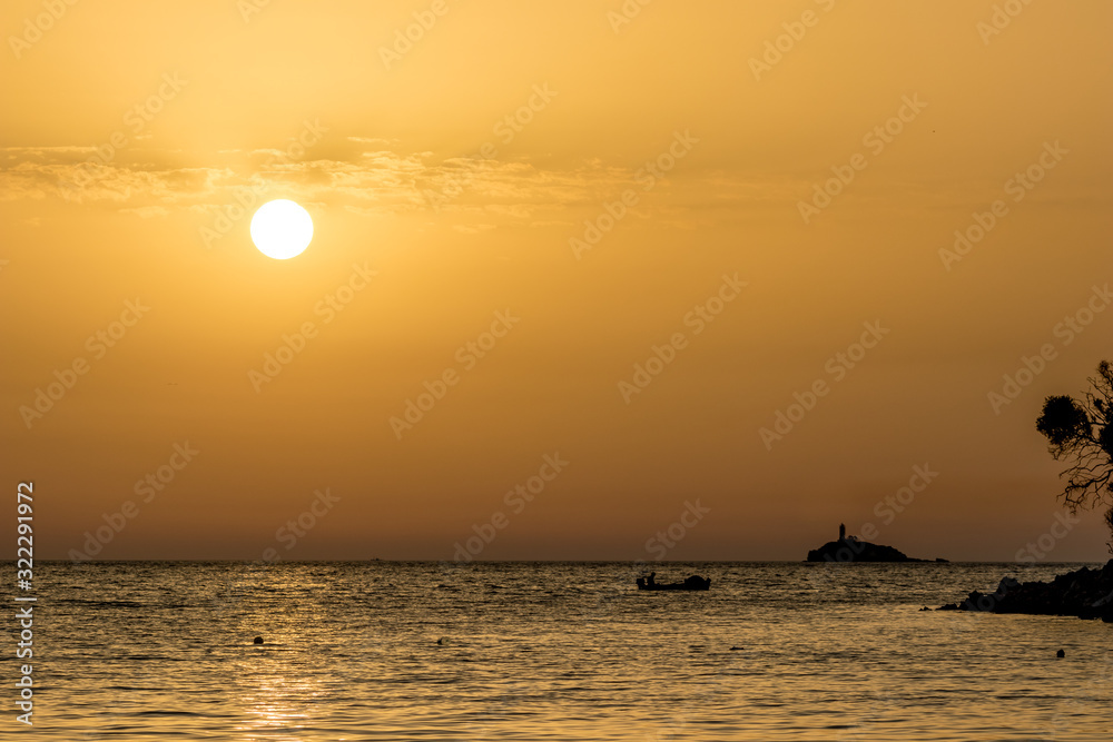 Focus on round setting Sun. Boat silhouette. Sun circle at beach at sunset in front of golden Ionian Sea water with slight overcast clouds. Dusk as seen from Ksamil, Albania, spring scenery evening Focus on round setting Sun. Boat silhouette. Sun circle at beach at sunset in front of golden Ionian Sea water with slight overcast clouds. Dusk as seen from Ksamil, Albania, spring scenery evening