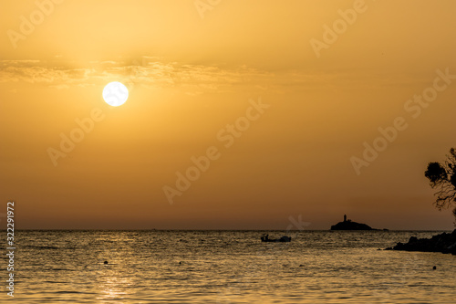 Focus on round setting Sun. Boat silhouette. Sun circle at beach at sunset in front of golden Ionian Sea water with slight overcast clouds. Dusk as seen from Ksamil  Albania  spring scenery evening