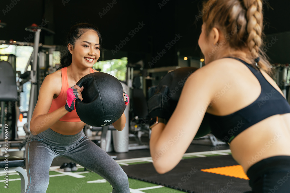 Fototapeta premium Happy young female athletic people performing squat exercises with friend and holding a medicine ball at fitness gym. Group of two confident women with healthy lifestyle working out together