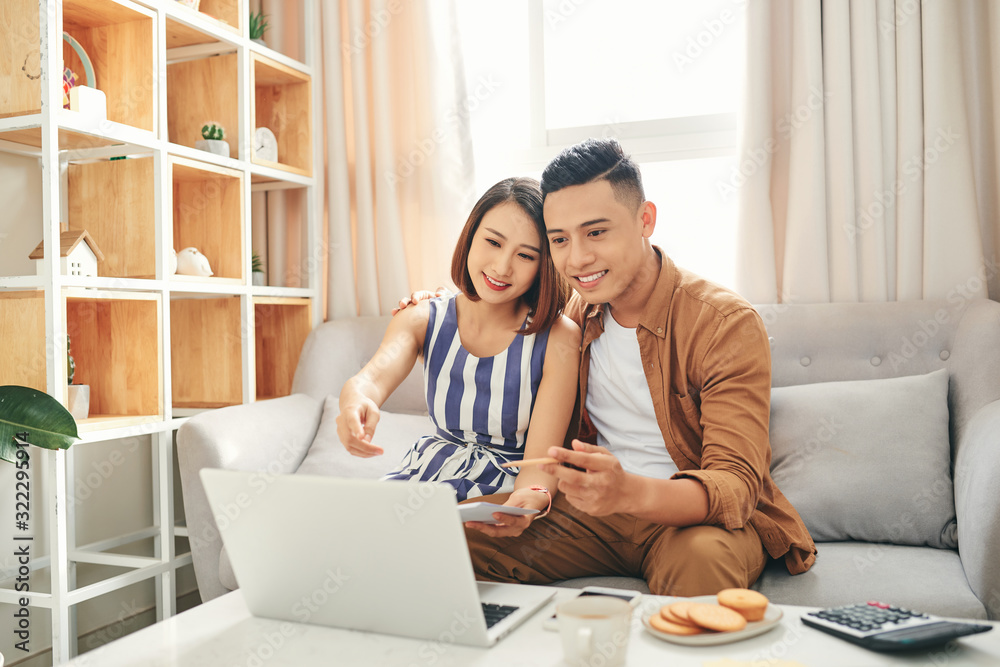 Smiling young couple sit on couch using laptop taking care of utility bills and house, read paperwork Smiling young couple sit on couch using laptop taking care of utility bills and house, read paperwork
