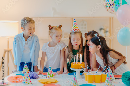 Small kids celebrate birthday party, blow candles on cake, gather at festive table, have good mood, enoy spending time together, make wish, wear party hats, pose indoor with inflated balloons photo