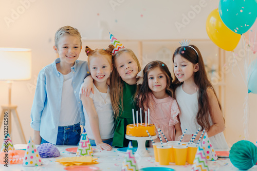 Horizontal shot of group of little children gather together to celebrate birthday, embrace and pose at camera, prepare for special occasion, stand near table with cake, paper cups, party caps
