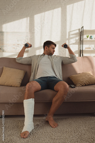 Handsome man with broken leg training with dumbbells on couch in living room