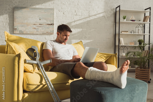 Canvas Print Handsome man with broken leg using laptop near crutches on sofa