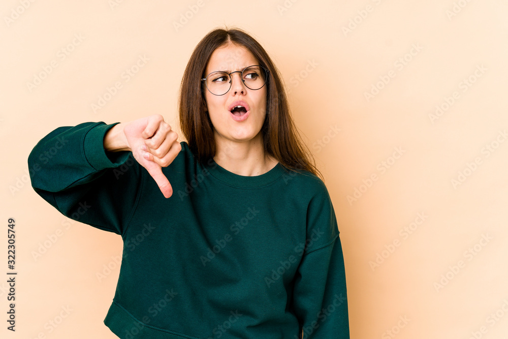 Young caucasian woman isolated on beige background showing thumb down and expressing dislike. Young caucasian woman isolated on beige background showing thumb down and expressing dislike.