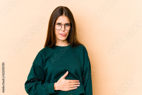 Young caucasian woman isolated on beige background touches tummy, smiles gently, eating and satisfaction concept. © Asier