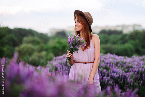 Red-haired girl in a hat lies in the grass with purple flowers. Young woman smile in nature. lady walks on a lavender field.